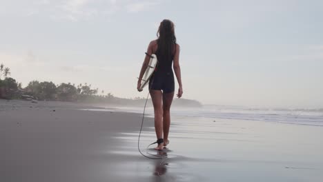 Vista-Trasera-De-Una-Mujer-Deportiva-Caminando-En-Una-Playa-De-Arena-Con-Su-Tabla-De-Surf