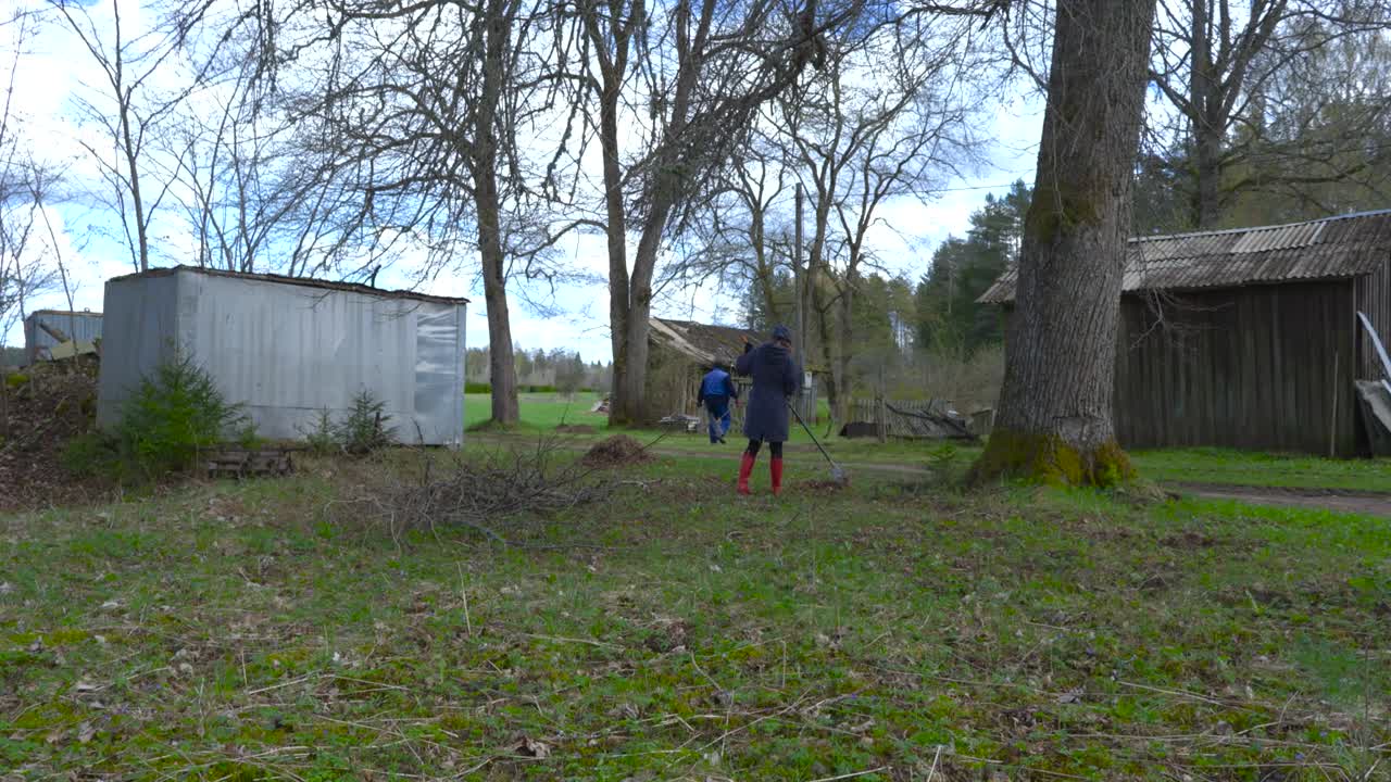 Timelapse footage of young energetic people raking leaves in a rural countryside garden when sun is occasionally coming out behind the clouds and lighting the green meadows and leaves on the ground.