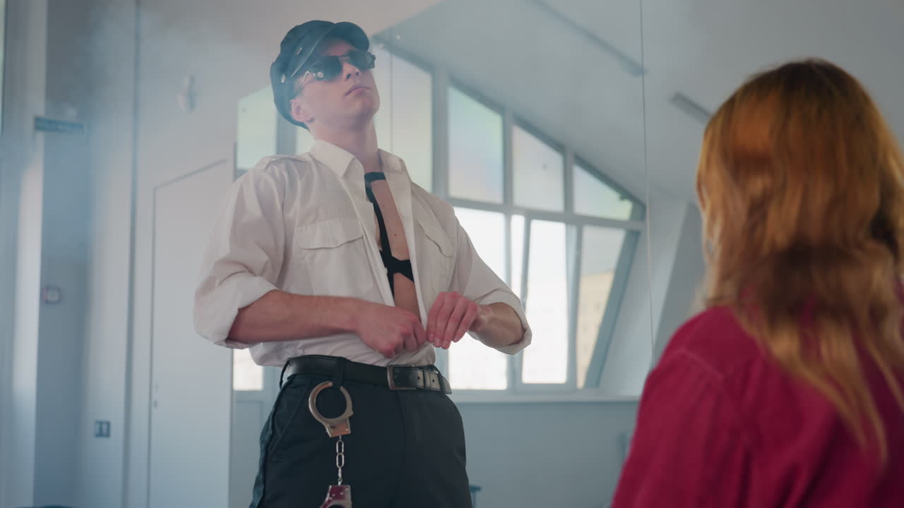 young man wearing cap and sunglasses removes shirt and flings it aside as he approaches seated woman amid smoke filling mirrored background and walls in bright minimalist studio room