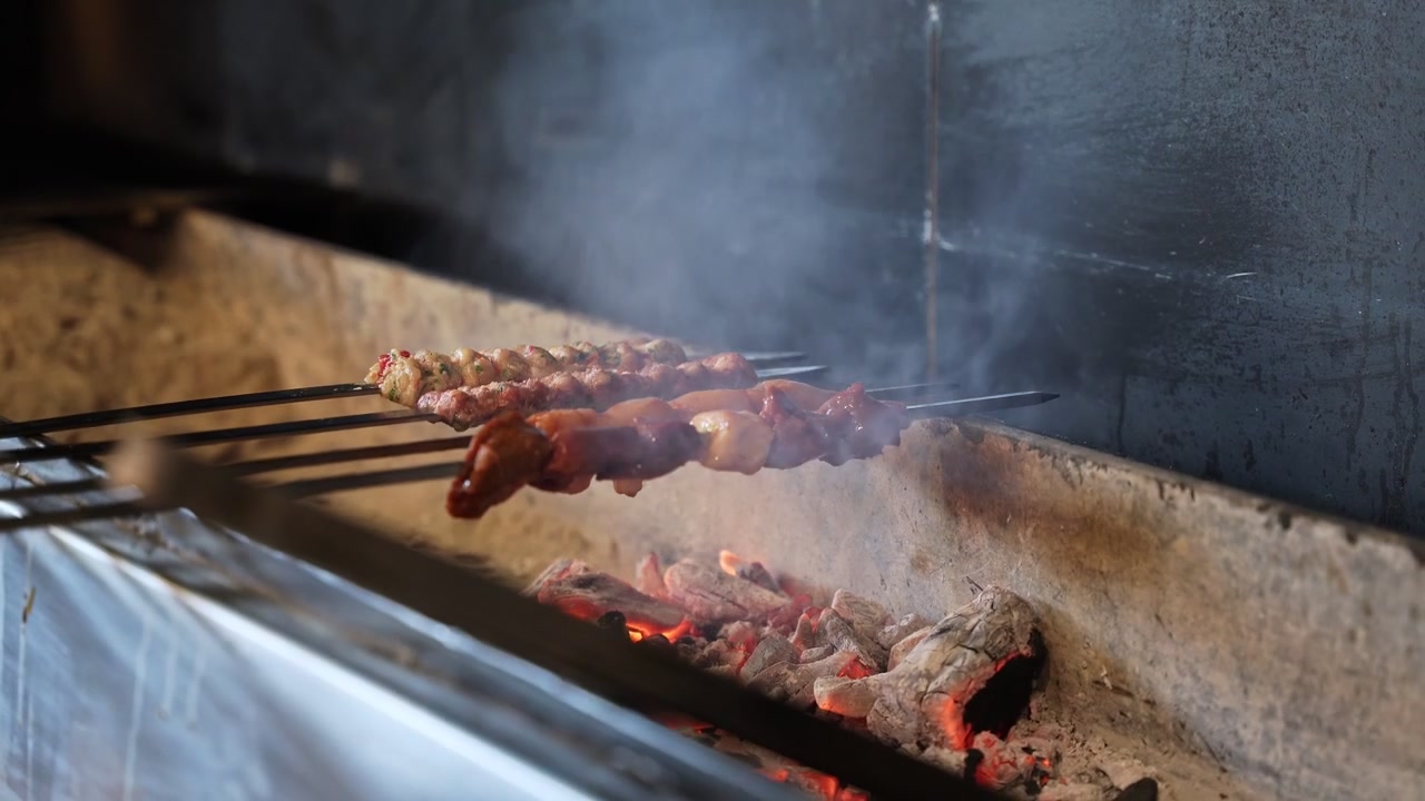 Un primo piano di spiedini tradizionali di carne e pollo che arrostiscono su una fossa di carbone ardente e fumante. I deliziosi kebab mediorientali cuociono lentamente su aste di metallo in un ambiente di cucina scuro e suggestivo.