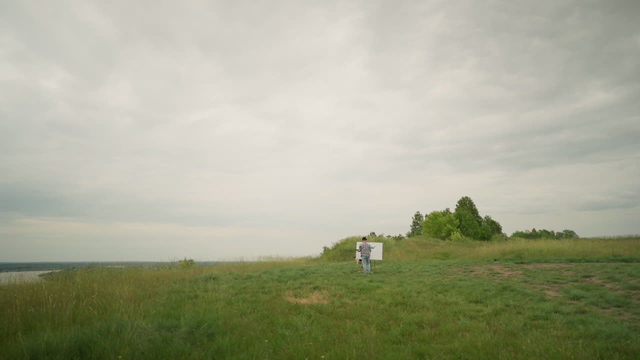 una vista trasera de un artista, con un sombrero y una camisa a cuadros, concentrado en pintar en un caballete en un campo verde y exuberante bajo un cielo nublado. la esencia de la creatividad al aire libre la soledad en la naturaleza