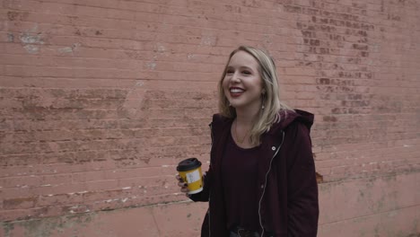 young blonde female smiling and walking down the street with a coffee in hand during the day