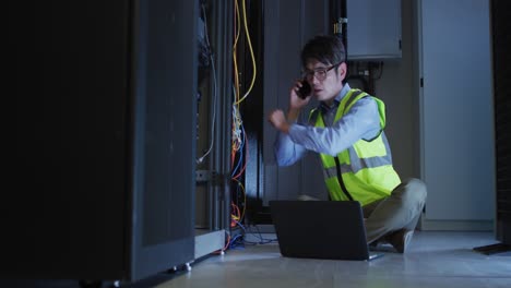 Asian-male-it-technician-using-smartphone-and-laptop-checking-computer-server