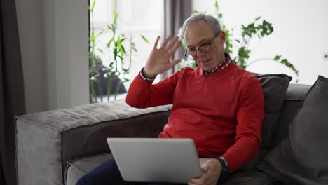 Un-Hombre-Mayor-Sonriente-Saluda-A-La-Cámara-Con-Una-Videollamada-En-Una-Computadora-Portátil