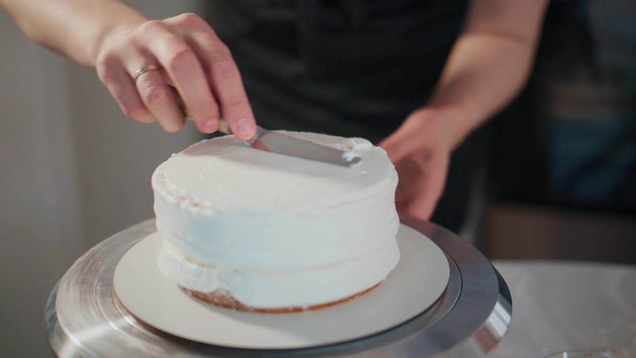 Close-up of hands smoothing white frosting on a cake. Cake spins slowly on a metal turntable. Person wears dark apron, focused on perfecting edges. Ideal for culinary, baking