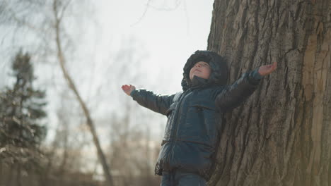 A young boy leans his back against a large tree trunk with his arms outstretched, looking upward, he is wearing a shiny black jacket, with trees and a blurred natural environment