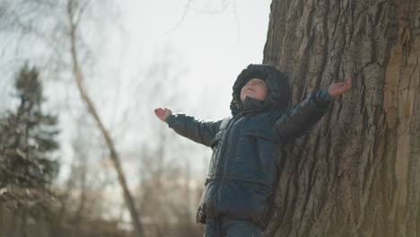 a young boy leans his back against a large tree trunk with his arms outstretched, looking upward, he is wearing a shiny black jacket, with trees and a blurred natural environment