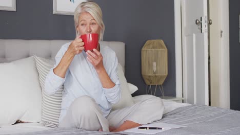 Happy-senior-caucasian-woman-sitting-on-bed-in-bedroom,-drinking-coffee