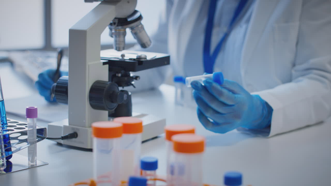 primer plano de un trabajador de laboratorio que realiza investigaciones utilizando un microscopio que sostiene un tubo de ensayo etiquetado lota