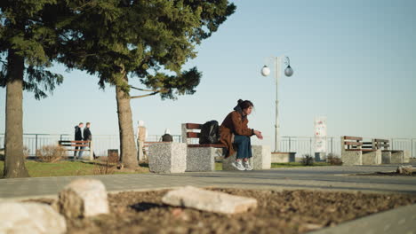 a girl wearing a brown coat, blue jeans, and white shoes sits alone on a bench in an urban park, her black backpack beside her. she looks downhearted and pensive, lost in thought