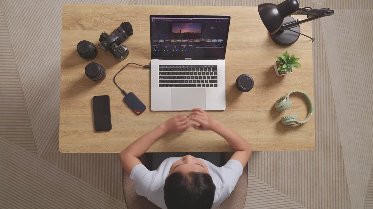 Top View Of Asian Woman Color Grading Thinking About The Footage While Sitting In The Workspace Using A Laptop Next To The Camera Editing The Video At Home