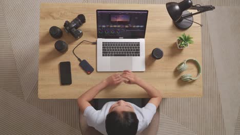 top view of asian woman color grading thinking about the footage while sitting in the workspace using a laptop next to the camera editing the video at home