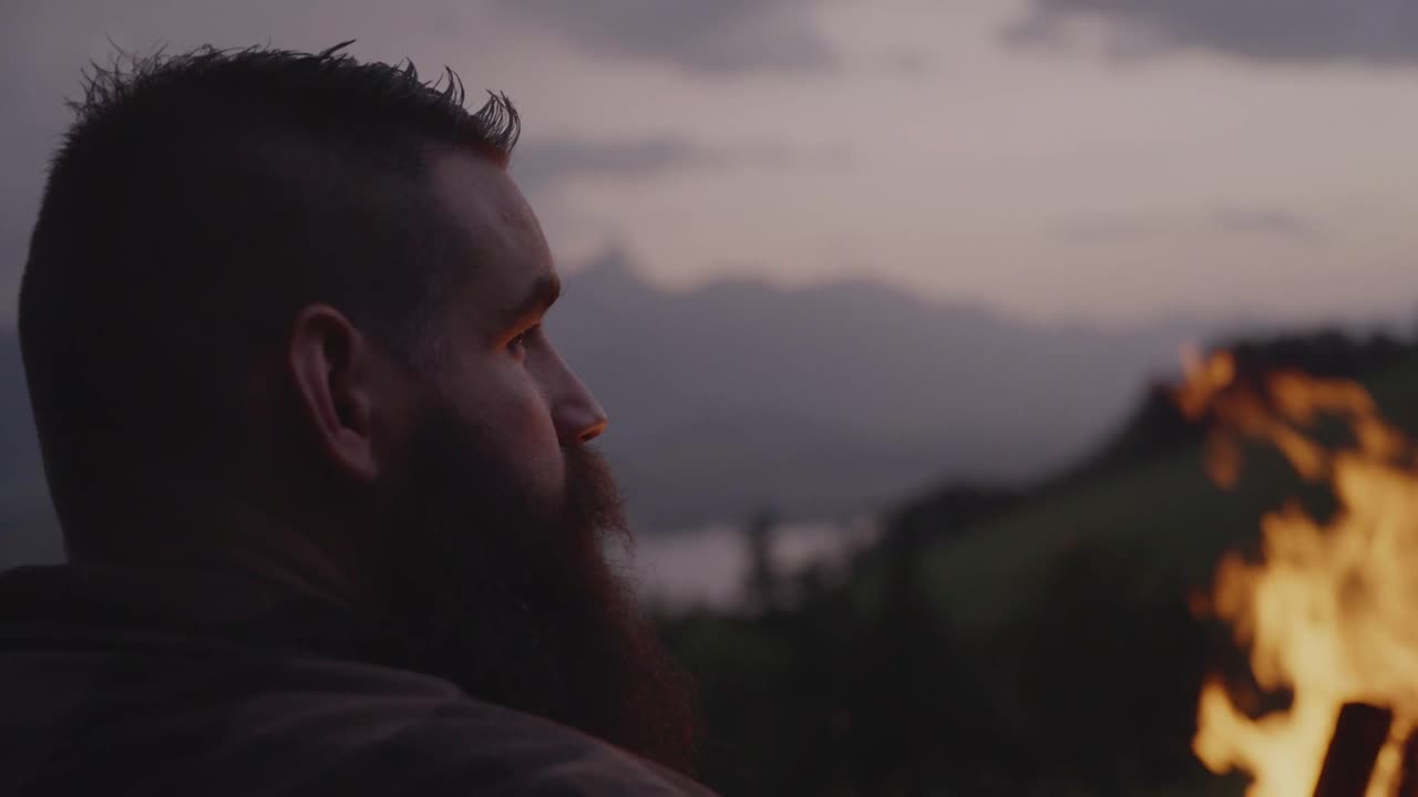 A man sits beside a glowing campfire, gazing toward the mountains during sunset. Peaceful outdoor moment symbolizing solitude, reflection, and connection with nature
