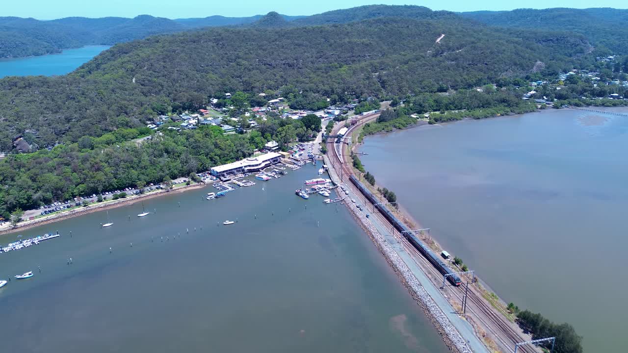 Paesaggio aereo con drone di un treno pendolare che si avvicina alla stazione ferroviaria di Hawkesbury River, vicino al sobborgo rurale di Brooklyn, circondato da boscaglia, con barche ormeggiate al molo sulla riva, in Australia.