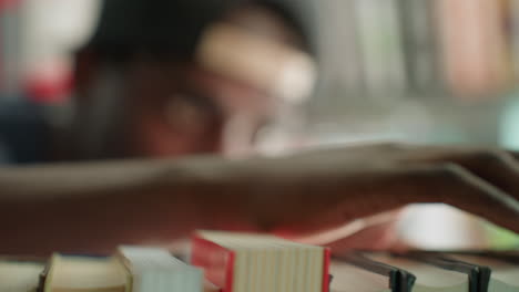 Primer plano de una mano que repasa libros en una estantería de una biblioteca, con un estudiante ligeramente desenfocado al fondo, poca profundidad de campo con tonos verde azulado, exploración táctil y selección cuidadosa del título
