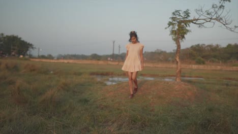 Slow-motion-wide-shot-of-a-pretty-indian-woman-dressed-in-a-pink-dress-walking-across-a-field-of-green-grass-while-walking-towards-the-camera-and-looking-thoughtful