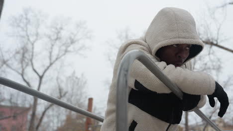 vista lateral de un hombre con suéter blanco descansando en barras de hierro, respirando pesadamente después de un intenso entrenamiento de invierno al aire libre, lleva guantes y una capucha, con una expresión enfocada en un parque nevado