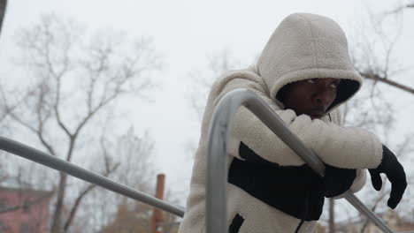 side view of man in white sweater resting on iron bars, breathing heavily after an intense outdoor winter workout, he is wearing gloves and a hood, with a focused expression in a snowy park setting