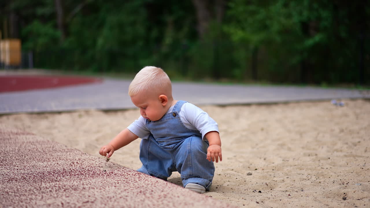 Little toddler boy wearing loose jeans romper pours sand from his hand. Child learning world playing outdoors.