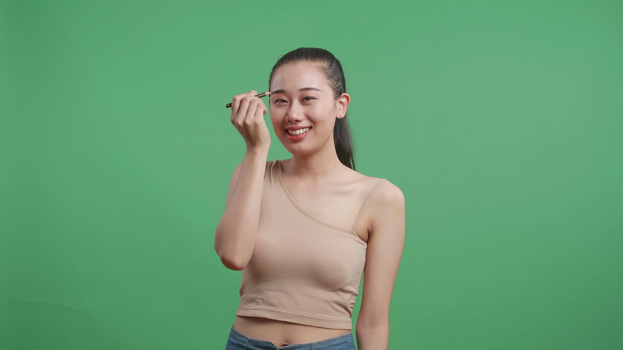 Asian Woman Applying Eyebrow Pencil And Smiling While Standing On Green Screen Background In The Studio
