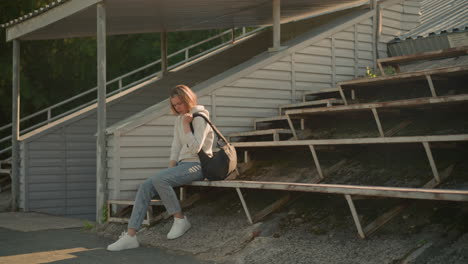 young woman with black bag adjusts her seating position on rusty stadium bleacher, stepping forward with right leg and repositioning bag