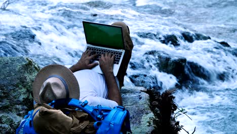 young man using green screen laptop computer on a waterfall. travel and freelance work concept