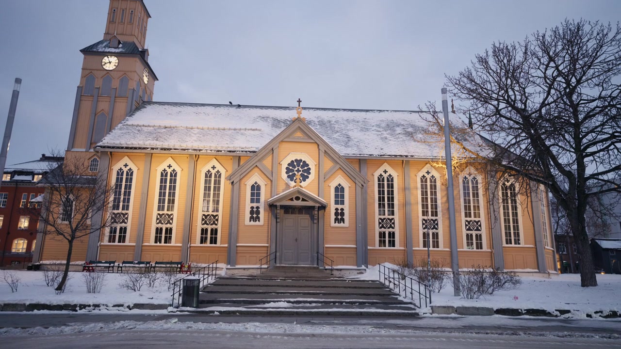 Tromssan tuomiokirkon yleiskuva edestä, lumen ympäröimänä talvisessa päivänvalossa, keskikokoinen staattinen kuva sisäänkäynnin portaista.