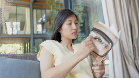 happy asian woman sitting on couch and reading book in sunny living room, slow motion