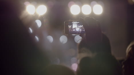 spectator man recording video of stage in front of bright spotlights via smartphone at outdoor music concert