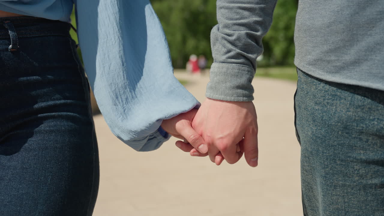 햇빛이 비치는 길에서 손을 잡고 있는 커플의 클로즈업, 데님 청바지와 파란색 소매가 보임, 부드럽게 잡은 손과 얽힌 손가락, 따뜻한 여름 햇살, 부드러운 연결을 전달하는 차분한 도시 공원 배경