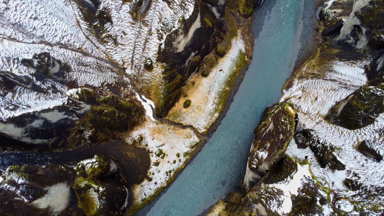 Aerial drone view of Iceland's Fjaorargljufur Canyon partially covered in snow, with a winding blue river cutting through