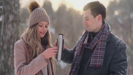 a loving couple man and woman in the winter forest drinking tea from a thermos. stylish man and woman in a coat in the park in winter for a walk. slow motion