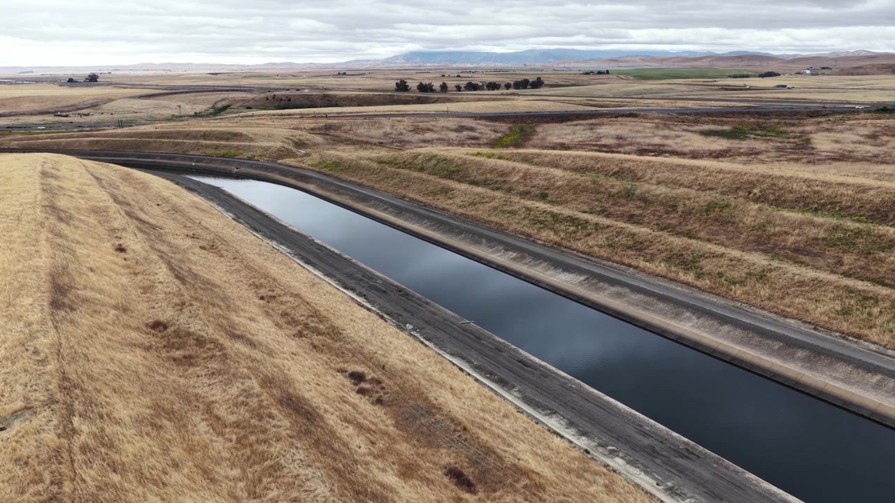 Acquedotto della California che si snoda attraverso aride colline dorate con una strada di accesso a lato, il fondo valle pianeggiante che si estende verso montagne distanti sotto un cielo pallido e coperto. California, USA. Ampia inquadratura aerea.