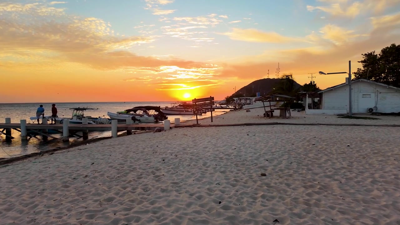 Pôr do sol tranquilo lançando um brilho quente sobre um cais de madeira com barcos atracados, capturando o fim sereno de um dia em ilha tropical. Ilha de Los Roques