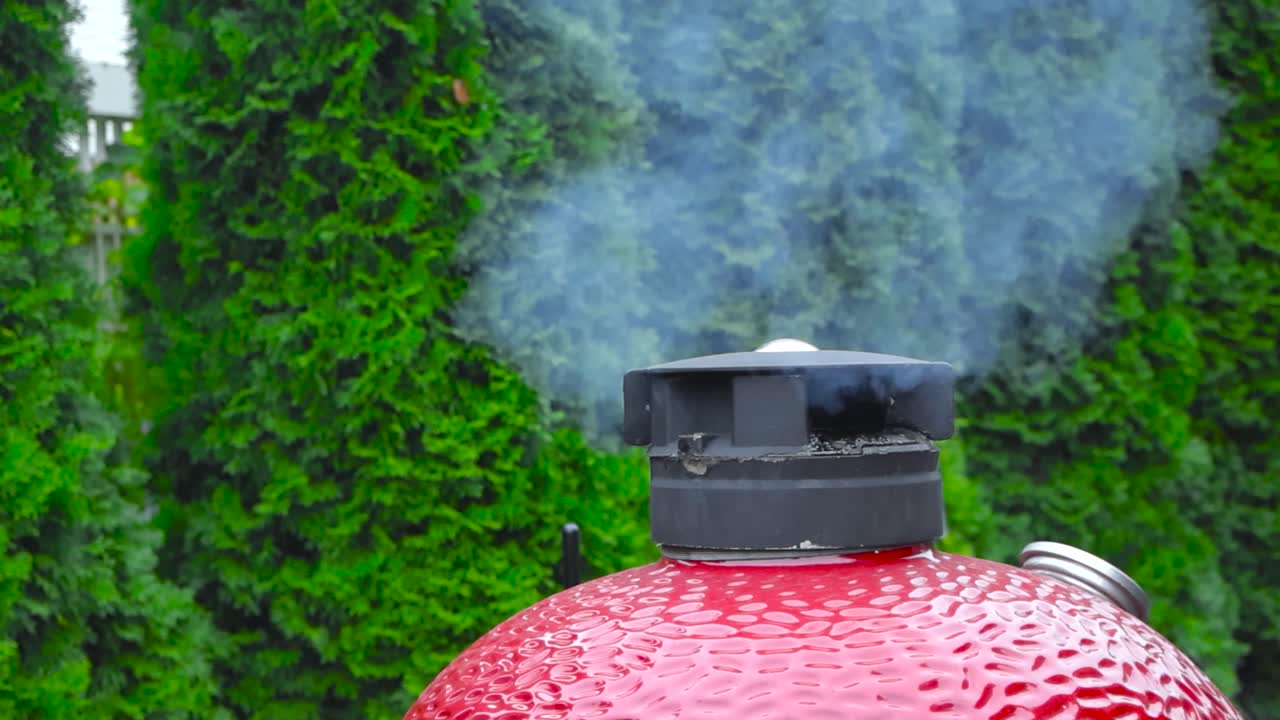 Griglia Komono rossa che fuma al rallentatore splendidamente durante il giorno in estate o autunno. Siepi o cespugli verdi sono sullo sfondo in una giornata o con tempo nuvoloso. Il termometro è visibile sulla griglia.