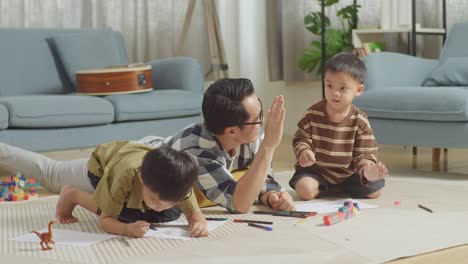 full body of asian father and sons on the floor in the room with plastic toy brick drawing together at home. clapping hands and giving high five celebrating on success drawing