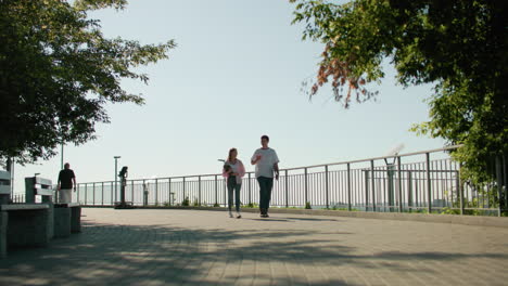 high school students walking holding books, engaging in cheerful conversation while a man passes by under bright sunlight with nearby bench, greenery, and urban architecture in background