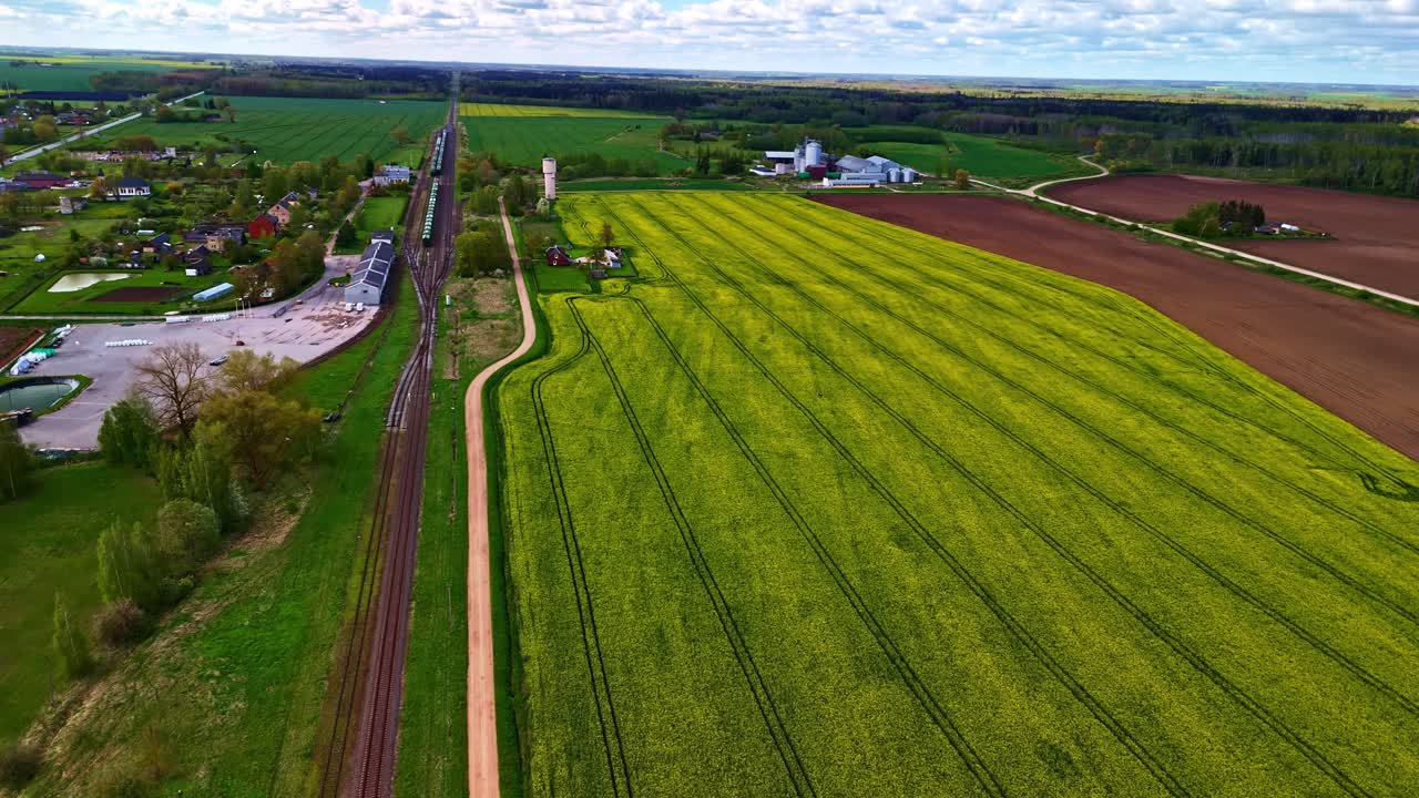Droneoptagelse over levende gule rapsmarker og grønne marker med et stillestående godstog langs en jernbanelinje nær en lille landsby og en gårdssilo.