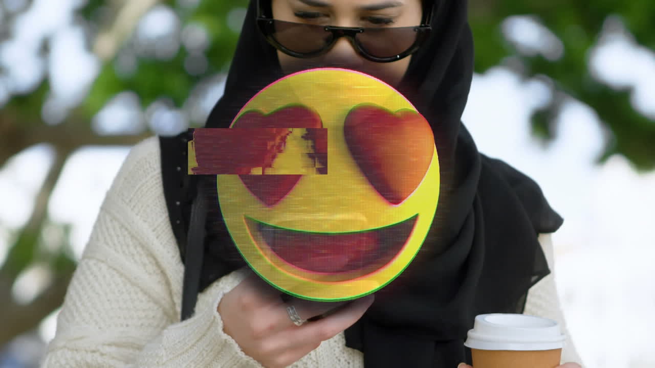 Animation of heart emoji over woman in hijab using smartphone