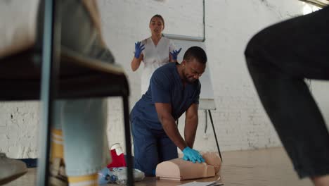 Bottom-view:-A-Black-male-doctor-performs-artificial-respiration-on-a-medical-mannequin,-and-his-assistant-girl-nurse-explains-to-the-public-against-a-white-brick-wall