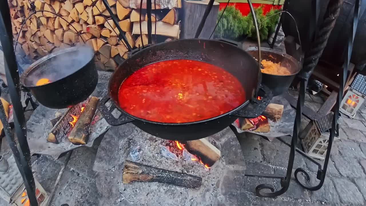 Tre calderoni bollono a fuoco vivo in una postazione di street food: zuppa rossa che sobbolle accanto a bollitori fumanti, con una catasta di legna e lanterne nelle vicinanze