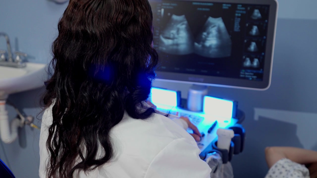Panning shot of a female doctor performing an abdominal ultrasound on a young Latina woman lying on an examination table in a modern medical clinic with blue LED lighting.