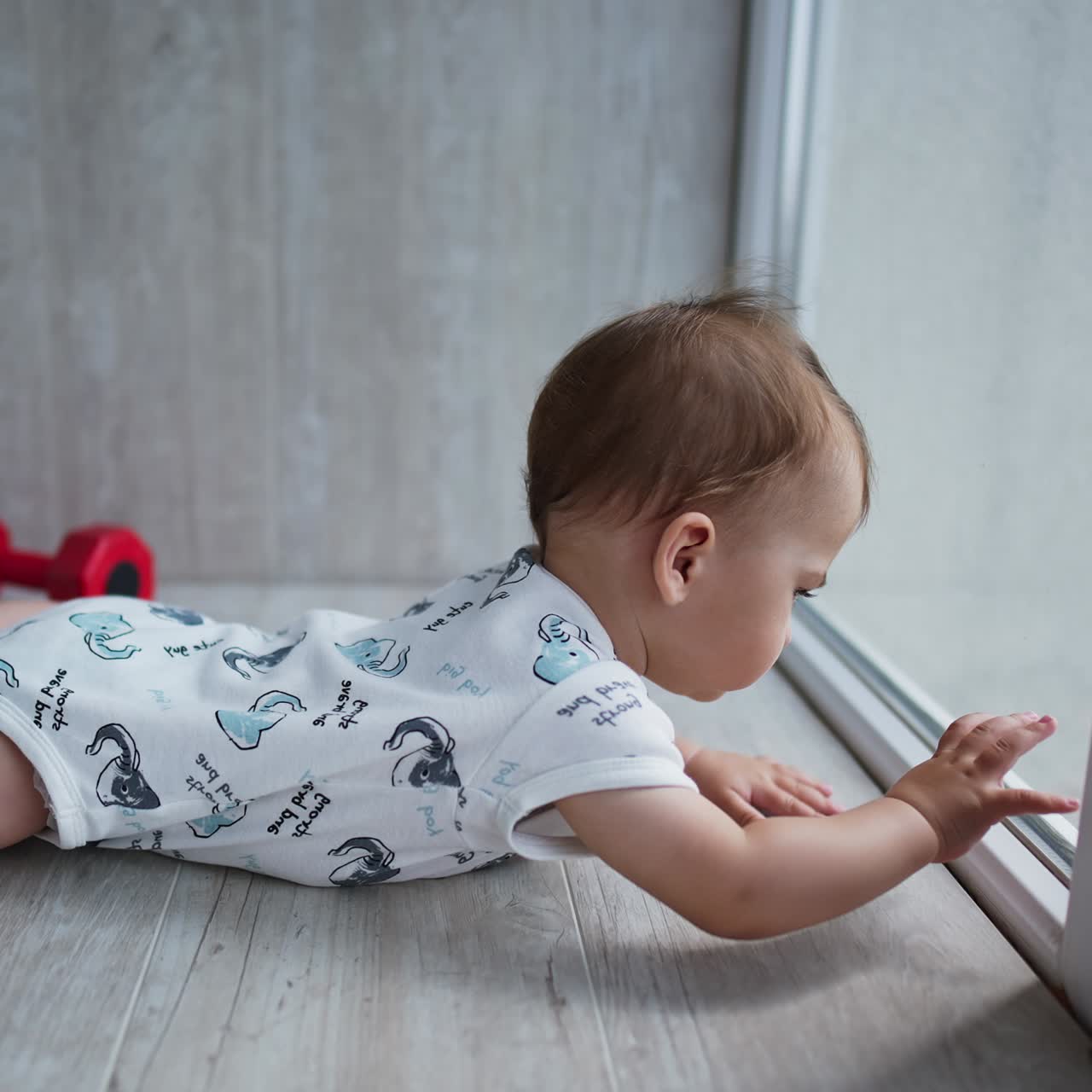 Little cute child on the wooden floor near the glass door. Adorable toddler touching the glass with his hands. Side view