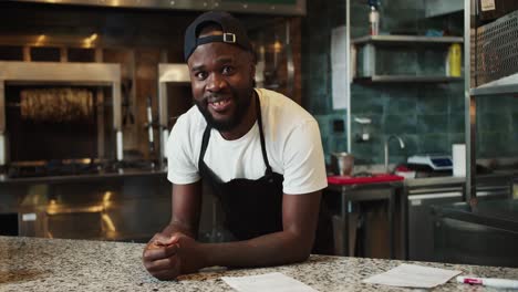 the chef in the doner market in a black apron is leaning on the tabletop, posing and looking at the camera. smiling doner cafe employee