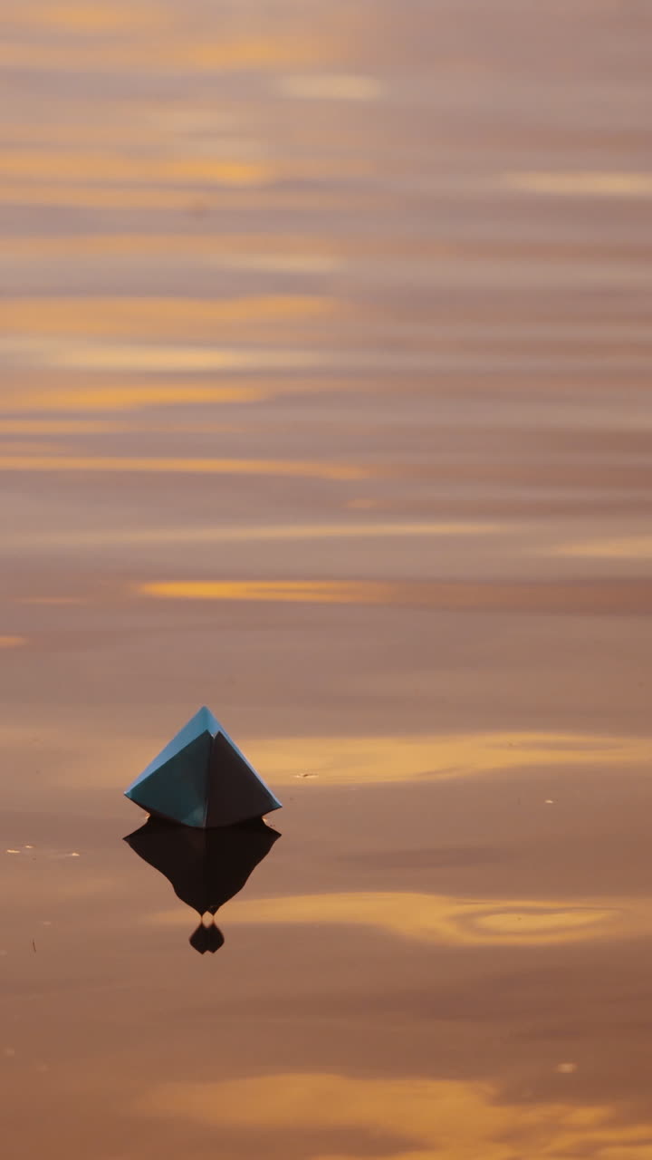 Paper boat on evening water. White origami boat floating alone on light waves on water surface at sunset. Vertical video