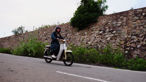 young caucasian woman with overcoat riding white moped motor scooter on countryside road