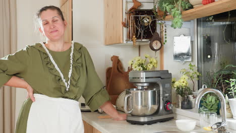 house wife in green dress and apron stands beside electric stand mixer in cozy kitchen filled with plants and wooden utensils, waiting for mixing process to finish while smiling warmly at camera