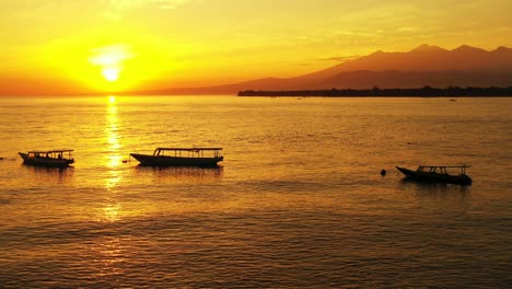 Silueta-De-Barcos-Flotando-En-La-Superficie-Del-Mar-En-Calma-Bajo-La-Puesta-De-Sol-Dorada-Con-El-Sol-Amarillo-Poniéndose-En-El-Horizonte-De-Las-Montañas-En-Indonesia