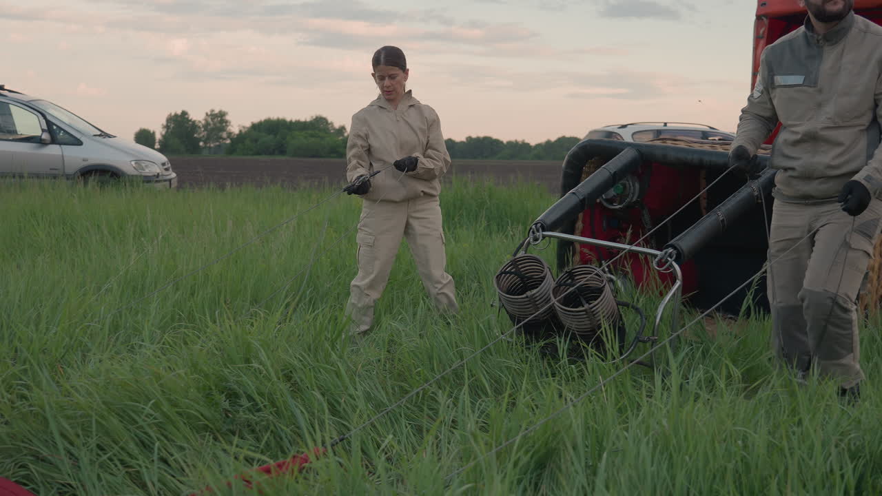 woman steps out from basket frame as man lowers burner assembly gradually against backdrop of deflating balloon envelope at dusk field crew preparing for flight and secure rope lines landing