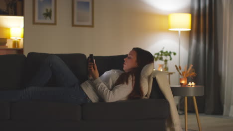 woman lying on sofa at home at streaming or watching movie or show or scrolling internet on mobile phone